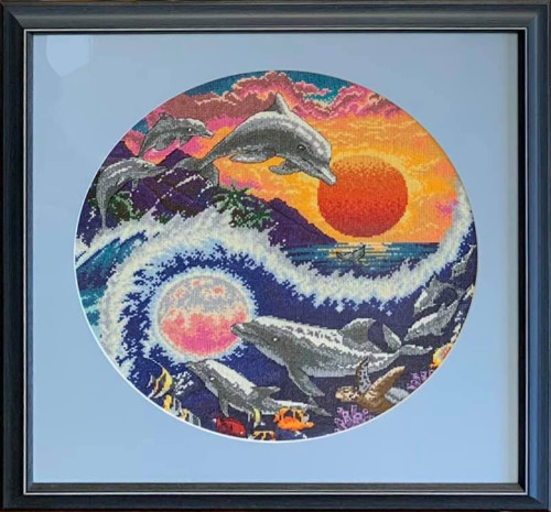 framed cross-stitch image depicting dolphins jumping out of sea with sun in background